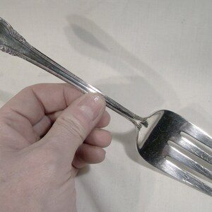 May include: A silver fork with a decorative handle. The fork has four tines and is held in a hand.