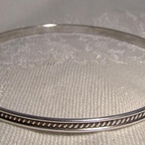 K&ouml;nnte beinhalten: Ein silbernes Armreif-Armband mit dekorativem Rand. Das Armband hat ein schlichtes, elegantes Design, perfekt f&uuml;r den Alltag. Der Rand weist ein sich wiederholendes Muster aus kleinen, dunklen Punkten und Linien auf. Das Armband wird auf einer wei&szlig;en, strukturierten Oberfl&auml;che gezeigt.