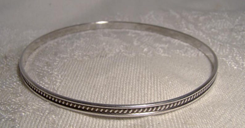 K&ouml;nnte beinhalten: Ein silbernes Armreif-Armband mit dekorativem Rand. Das Armband hat ein schlichtes, elegantes Design, perfekt f&uuml;r den Alltag. Der Rand weist ein sich wiederholendes Muster aus kleinen, dunklen Punkten und Linien auf. Das Armband wird auf einer wei&szlig;en, strukturierten Oberfl&auml;che gezeigt.