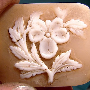May include: A white cameo carved with a delicate flower and leaves on a light brown background.