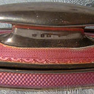 May include: A silver and pink enamel compact with a hinged lid. The lid has a raised, oval-shaped design. The compact is decorated with a delicate, repeating pattern.