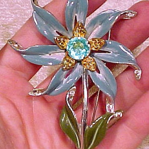 May include: A blue enamel flower brooch with a light blue rhinestone centre and gold accents. The flower has five petals and a stem with green leaves.