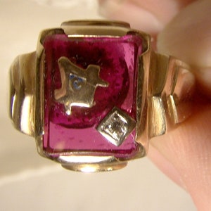 May include: A gold ring with a square, faceted, dark red stone. The stone has a gold design inside.
