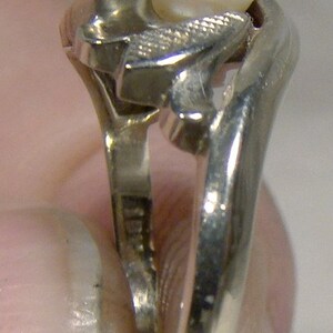 May include: A silver ring with a pearl set in a stylized design. The ring is worn on a finger.