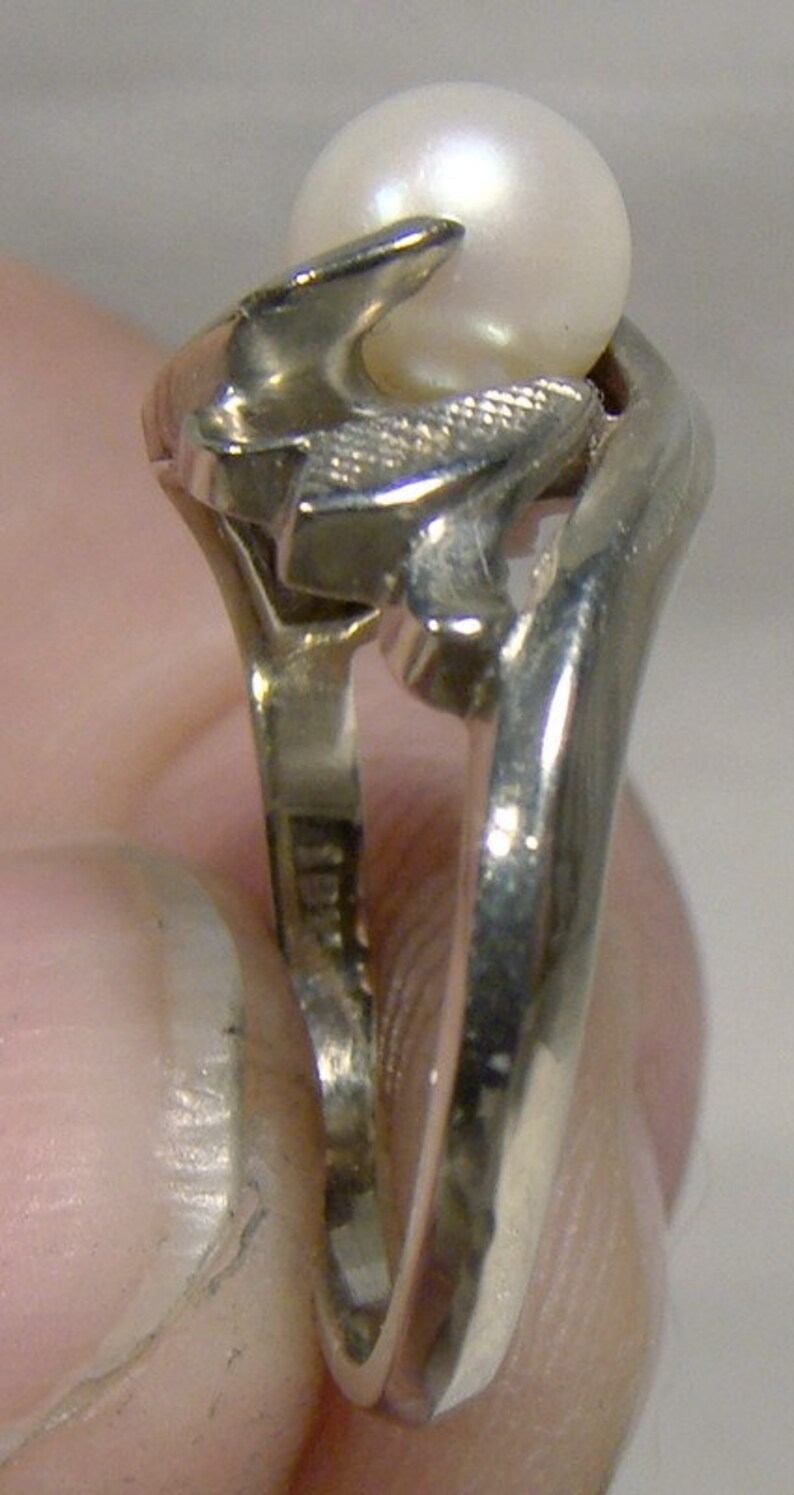 May include: A silver ring with a pearl set in a stylized design. The ring is worn on a finger.