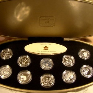 May include: A gold-colored oval-shaped box with a hinged lid, containing a set of 12 silver coins. The coins are arranged in a circle around a small card with the text "Millennium Coins - Pièces de monnaie du millénaire".