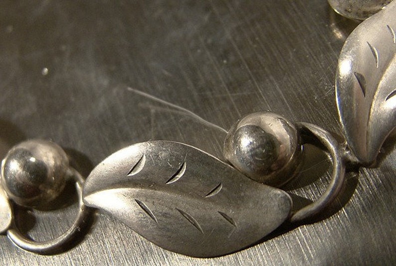 K&ouml;nnte beinhalten: Ein silbernes Armband mit blattf&ouml;rmigen Anh&auml;ngern und runden Perlen. Die Bl&auml;tter haben eine strukturierte Oberfl&auml;che und sind durch silberne Ringe miteinander verbunden.