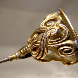 May include: A gold-toned metal hat pin with a decorative, ornate design. The pin has a long, thin stem and a large, intricate head.
