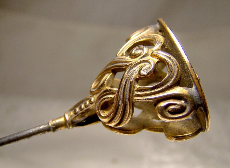 May include: A gold-toned metal hat pin with a decorative, ornate design. The pin has a long, thin stem and a large, intricate head.