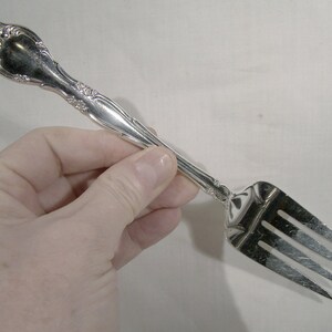 May include: A silver fork with an ornate handle. The fork has three tines and is held in a hand.