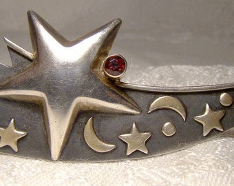 May include: A silver brooch in the shape of a shooting star with a red gemstone. The star is surrounded by smaller stars and crescent moons.