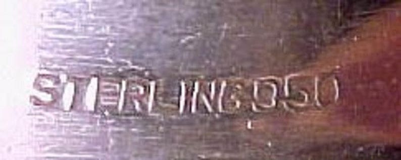 Pu&ograve; includere: Primo piano di un oggetto d'argento con l'iscrizione "STERLING 950" incisa sulla superficie.