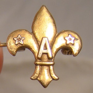 May include: A gold-toned fleur-de-lis badge with the letter "A" in the centre. The badge features two stars, one on each side. The design suggests a vintage or antique item, possibly a pin or emblem.
