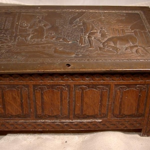 May include: A wooden box with a carved design and a brass lid featuring an embossed scene of people and animals. The box has a hinged lid and a decorative design on the front.