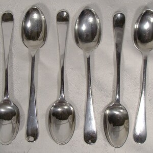 May include: Six silver spoons with rounded bowls and simple handles. The spoons are arranged in a row on a white surface.