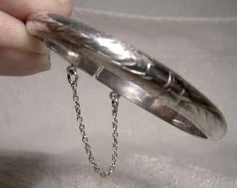 May include: A silver bangle bracelet with a delicate chain attached. The bracelet has a textured design and is open on one side.
