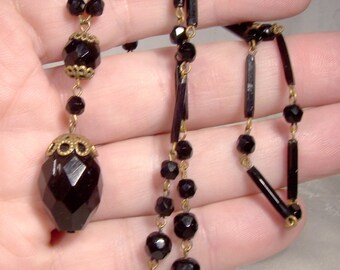 Könnte beinhalten: Eine lange schwarze Halskette mit einem großen, facettierten schwarzen Glasanhänger. Die Halskette besteht aus schwarzen Glasperlen und einer goldenen Kette.