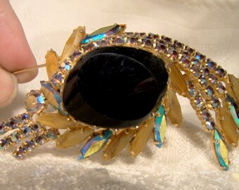 Könnte beinhalten: Eine goldfarbene Brosche mit einem großen schwarzen Cabochonstein, umgeben von schillernden Strasssteinen und goldfarbenen Blättern. Die Brosche hat einen herabhängenden Fransen aus schillernden Strasssteinen.