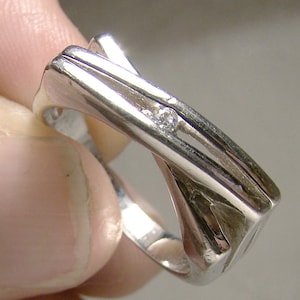 May include: A silver ring featuring a single, small, clear diamond set in the centre. The ring has a distinctive, modern design with a criss-cross pattern.