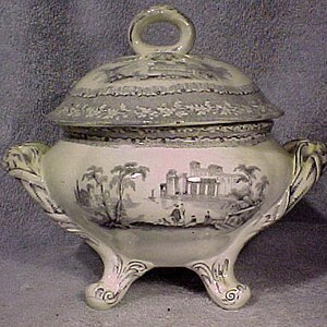 May include: A white porcelain tureen with a lid, featuring a grey and white floral design and a scene of a building with trees. The tureen has three legs and two handles.