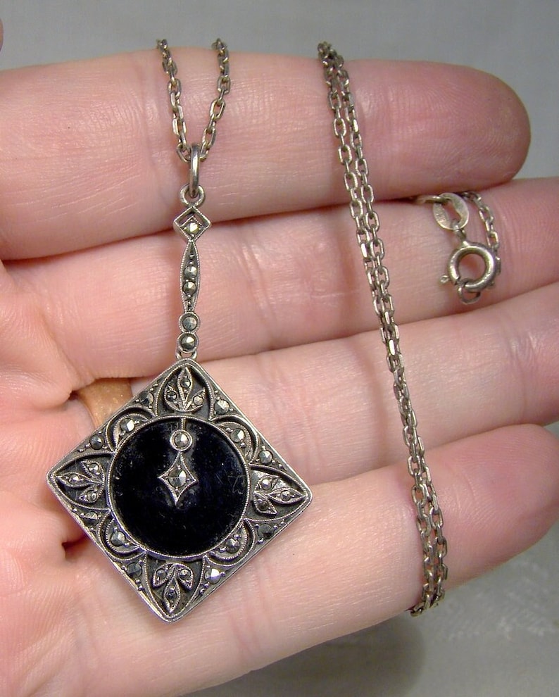 May include: A silver necklace with a black onyx pendant. The pendant is square shaped and features a detailed design of marcasite stones. The chain is a delicate silver chain.