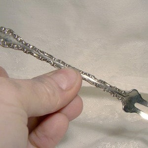 May include: A silver ornate fork with a decorative handle. The fork is held in a hand against a white background.