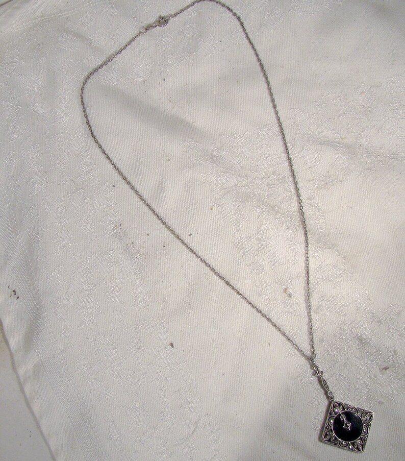 May include: A silver chain necklace with a black and silver pendant. The pendant is a square with a black stone in the center and silver filigree around the edges.
