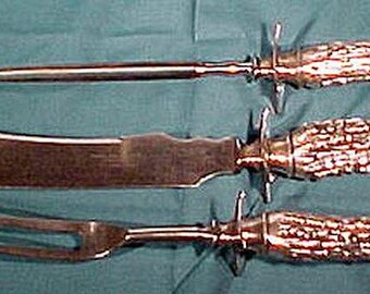 Könnte beinhalten: Ein dreiteiliges Tranchierbesteck mit Hirschhorn-Griffen. Das Set enthält ein Tranchiermesser, eine Tranchiergabel und einen Wetzstahl. Die Klingen und Zinken sind silberfarben, die Griffe hellbraun.