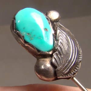 May include: A silver and turquoise pin with a teardrop-shaped turquoise stone. The pin has a detailed design with a silver wing and a round silver accent.