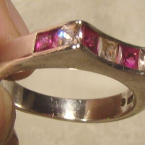 May include: A silver ring with a V-shaped top, set with alternating pink and clear square-cut gemstones. The ring is held by a person's fingers, showcasing its design and sparkle.