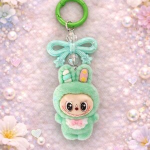 Puede incluir: Un llavero de conejito verde menta con un cierre verde y un lazo. El conejito de peluche tiene una cara sonriente, una pajarita rosa y detalles amarillos en las orejas. El fondo es rosa suave y brillante.