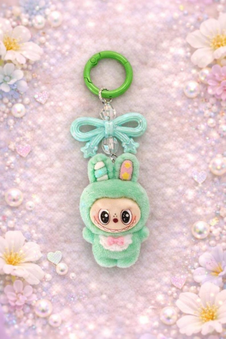 Puede incluir: Un llavero de conejito verde menta con un cierre verde y un lazo. El conejito de peluche tiene una cara sonriente, una pajarita rosa y detalles amarillos en las orejas. El fondo es rosa suave y brillante.