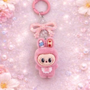 Puede incluir: Un llavero de conejito rosa con un lazo y cierre rosa a juego. El conejito de peluche lleva un traje con capucha con un babero blanco y una cara sonriente. El fondo es rosa suave con flores y corazones.