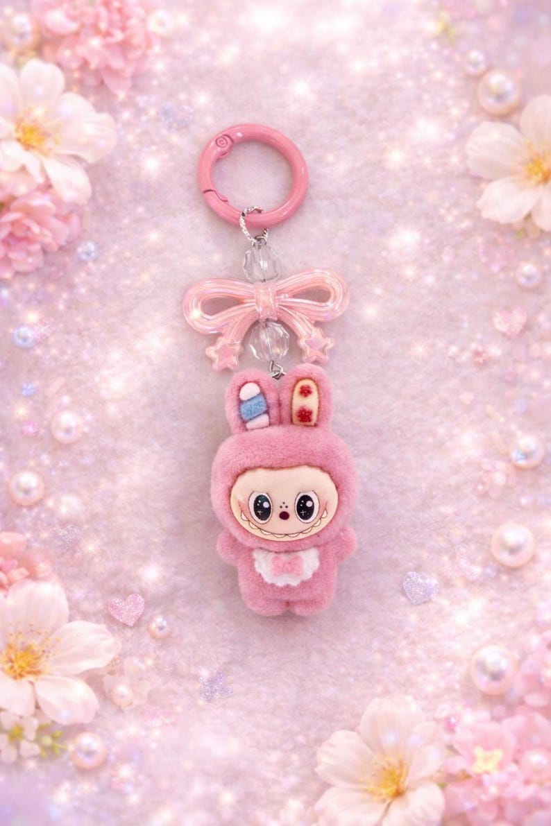 Puede incluir: Un llavero de conejito rosa con un lazo y cierre rosa a juego. El conejito de peluche lleva un traje con capucha con un babero blanco y una cara sonriente. El fondo es rosa suave con flores y corazones.