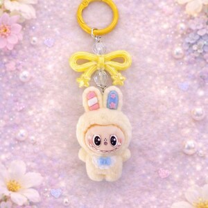 Puede incluir: Un llavero caprichoso con un personaje de conejito de peluche con un atuendo con capucha de color crema. El conejito tiene detalles en rosa y azul en las orejas y un babero azul. Un lazo y un anillo amarillos completan el dise&ntilde;o.