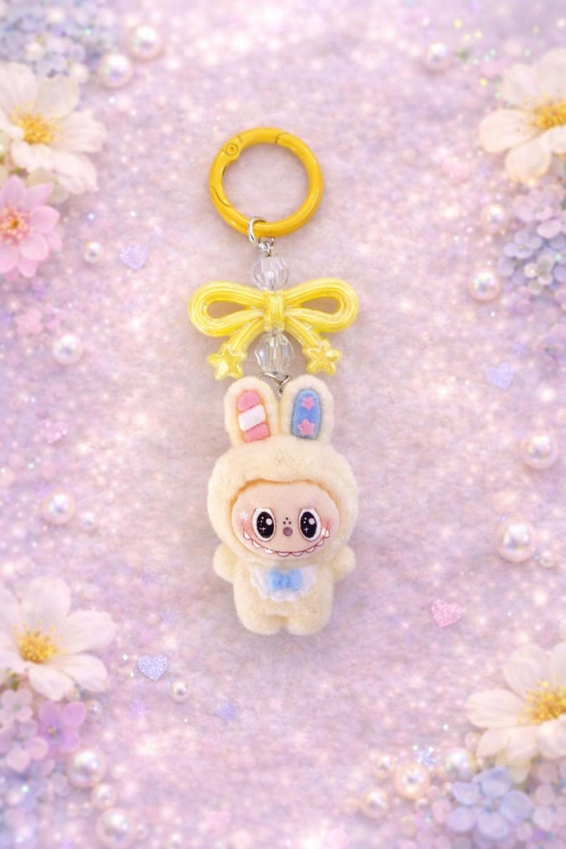 Puede incluir: Un llavero caprichoso con un personaje de conejito de peluche con un atuendo con capucha de color crema. El conejito tiene detalles en rosa y azul en las orejas y un babero azul. Un lazo y un anillo amarillos completan el dise&ntilde;o.
