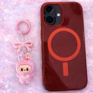 Puede incluir: Un llavero de conejito rosa con un lazo y una funda roja para tel&eacute;fono con un dise&ntilde;o circular. La funda y el llavero est&aacute;n sobre una superficie rosa brillante. El llavero de conejito tiene una cara linda.