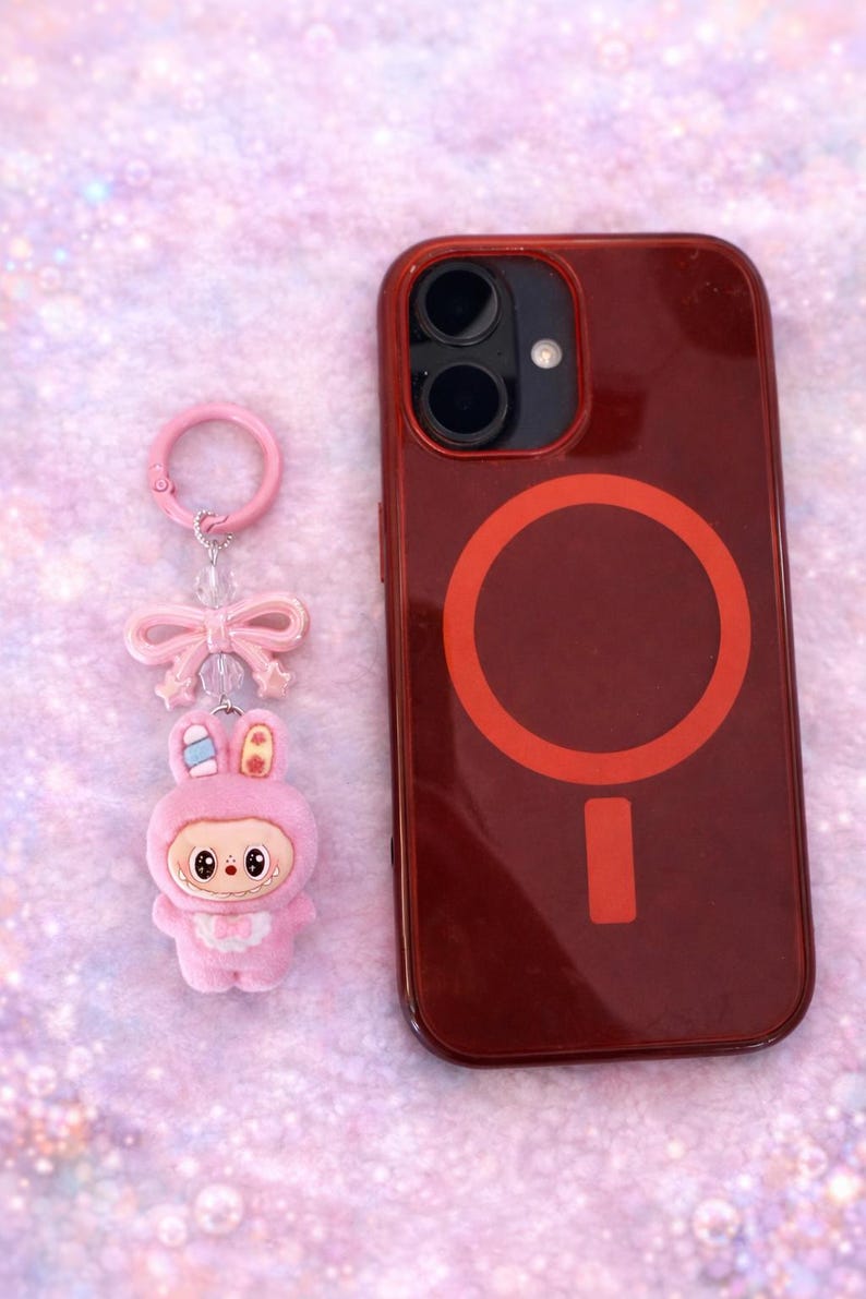 Puede incluir: Un llavero de conejito rosa con un lazo y una funda roja para tel&eacute;fono con un dise&ntilde;o circular. La funda y el llavero est&aacute;n sobre una superficie rosa brillante. El llavero de conejito tiene una cara linda.