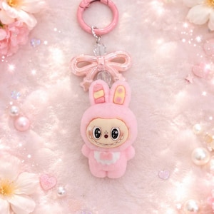 Puede incluir: Un llavero de conejo rosa con un dise&ntilde;o de dibujos animados. El conejo lleva un atuendo con capucha y tiene una cara sonriente. El llavero incluye un lazo rosa y un cierre rosa. El fondo es una textura suave y rosa con flores decorativas.