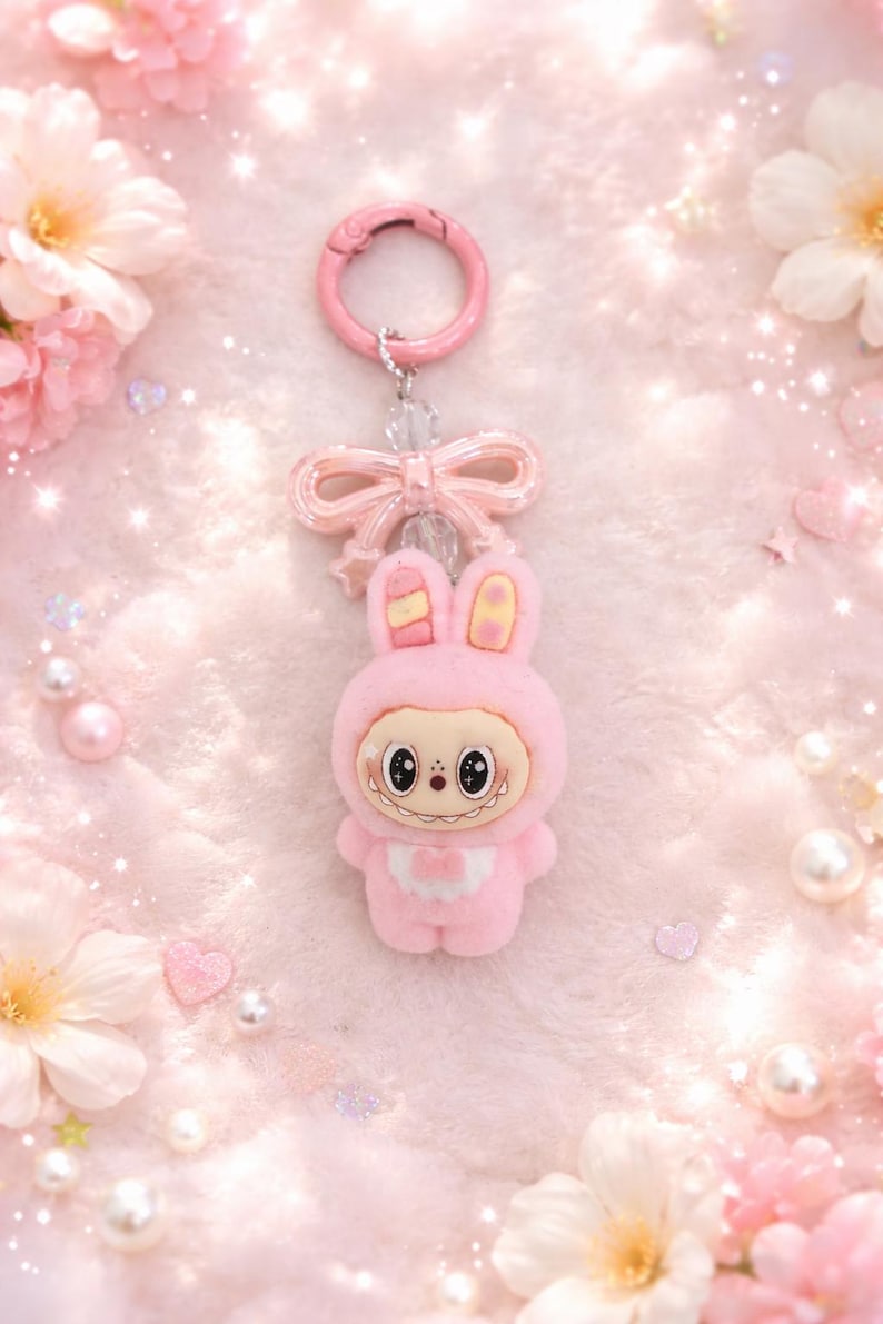 Puede incluir: Un llavero de conejo rosa con un dise&ntilde;o de dibujos animados. El conejo lleva un atuendo con capucha y tiene una cara sonriente. El llavero incluye un lazo rosa y un cierre rosa. El fondo es una textura suave y rosa con flores decorativas.