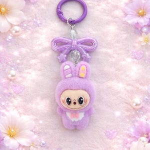Puede incluir: Un llavero de conejito lavanda con un lazo morado y un cierre redondo. El conejito tiene una cara linda, una pajarita rosa en forma de coraz&oacute;n y detalles amarillos en las orejas. El fondo es rosa suave con flores y perlas.