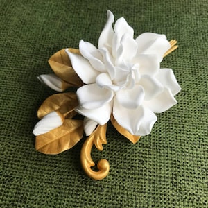 May include: A decorative brooch featuring a large, multi-petaled white gardenia flower with two smaller white buds. The floral arrangement is accented by several metallic gold leaves and a curved, ornate gold stem detail at the base.