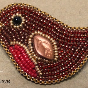 Op de afbeelding: Een broche in de vorm van een vogel, gemaakt van kralen. In het midden is een koperkleurig bladmotief te zien. De vogel is gemaakt van rode, gouden en zwarte kralen.