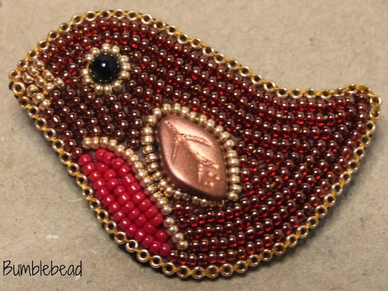 Op de afbeelding: Een broche in de vorm van een vogel, gemaakt van kralen. In het midden is een koperkleurig bladmotief te zien. De vogel is gemaakt van rode, gouden en zwarte kralen.