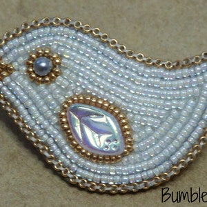 Op de afbeelding: Een witte kralen vogel broche met gouden accenten en een parelmoerachtig centrum. De vogel is omrand met een gouden ketting.