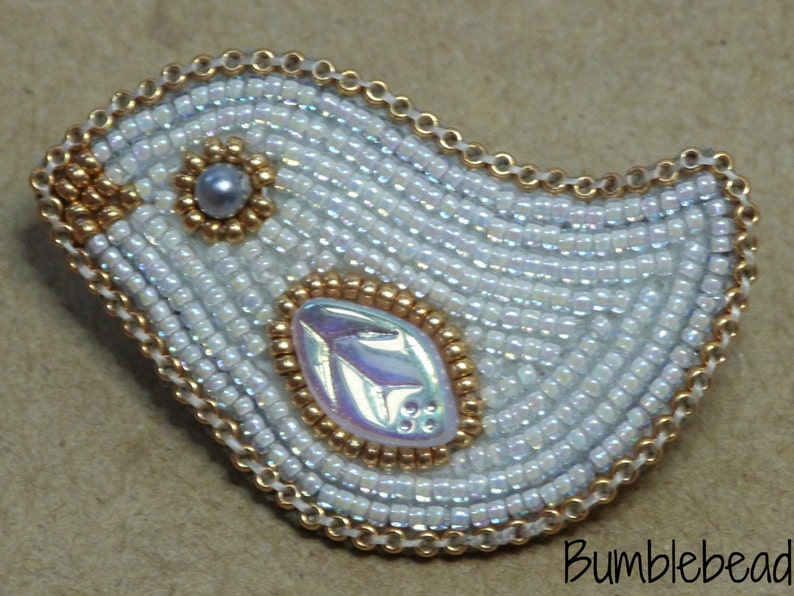 Op de afbeelding: Een witte kralen vogel broche met gouden accenten en een parelmoerachtig centrum. De vogel is omrand met een gouden ketting.