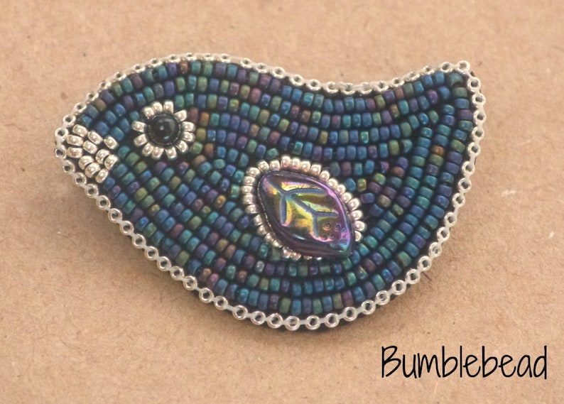 Op de afbeelding: Een geperst vogel broche met een iriserende blauwe, groene en paarse kleurstelling. De vogel heeft een zwart oog en een zilveren bladontwerp in het midden.