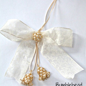 May include: A white satin ribbon bow with a gold and pearl embellishment and two pearl and gold bells hanging from the center.