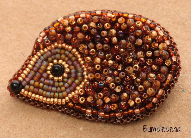 May include: A brown and gold beaded hedgehog brooch. The hedgehog is made of small, round beads in various shades of brown, gold, and iridescent colours. The eye is a black bead.