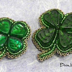 May include: Two green four-leaf clover brooches made with green glass beads and gold beads. The brooches are on a light purple background.